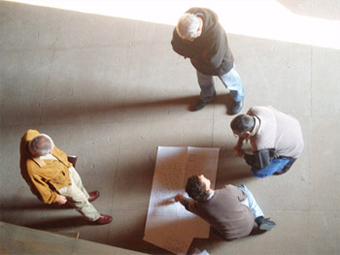 Foto di quattro persone riprese dall'alto. Due persone inginocchiate e due in piedi che stanno discutendo di un progetto cartaceo posato in terra 